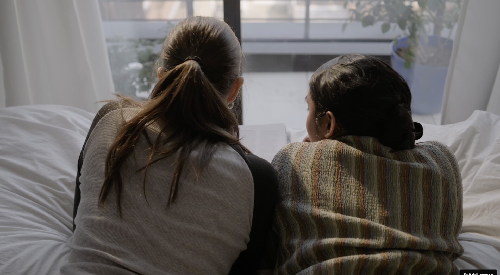 Two people on a bed look out a window with white curtains.